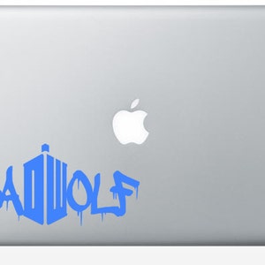 May include: A blue vinyl decal on a silver laptop. The decal features the word "BAD WOLF" in a graffiti-style font, with a stylized police box design. The Apple logo is visible.
