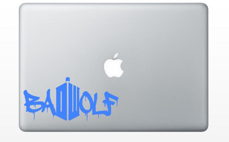May include: A blue vinyl decal on a silver laptop. The decal features the word "BAD WOLF" in a graffiti-style font, with a stylized police box design. The Apple logo is visible.