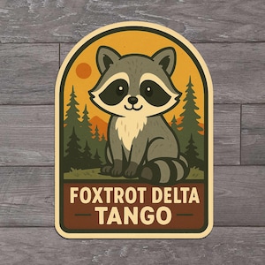 May include: A rectangular sticker featuring a cartoon raccoon in a forest scene with a setting sun. The raccoon is gray and white with black markings. The sticker has the text "FOXTROT DELTA TANGO" in a brown banner.