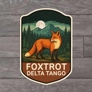 May include: A sticker featuring a stylized illustration of a fox in a forest scene under a full moon. The fox is orange and white, with a dark green forest and mountains in the background. The text "FOXTROT DELTA TANGO" is at the bottom.