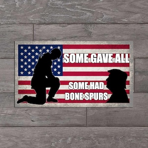 May include: Rectangular sign featuring the American flag with the text "SOME GAVE ALL" and "SOME HAD BONE SPURS." A kneeling soldier silhouette is on the left, and a silhouette of a man is on the right.