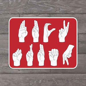 May include: A red rectangular sign featuring white illustrations of hands in various positions, representing the American Sign Language alphabet. The sign has a white border and is set against a wooden background.