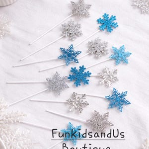 Könnte beinhalten: Eine Sammlung von schneeförmigen Dekorationen in Silber und blauem Glitzer, befestigt an weißen Stäben. Die Dekorationen sind auf einer weißen Oberfläche angeordnet, mit dem Text "FunkidsandUs Boutique".