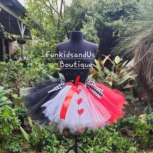 May include: A red, white and black tulle tutu with a checkerboard pattern and red hearts. The tutu is on a mannequin and has a red ribbon around the waist. The text "FunkidsandUs Boutique" is visible on the mannequin.