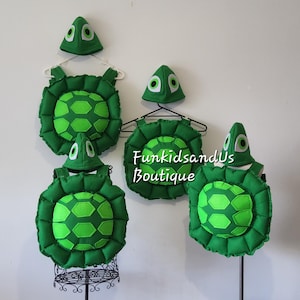 May include: Several green turtle costumes with matching hats are displayed. The costumes have a round, segmented shell design with a lighter green pattern in the center. The hats have white eye details. The text "FunkidsandUs Boutique" is visible.