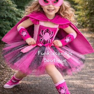 Op de afbeelding: Een roze en zwart superhelden kostuum voor kinderen. De outfit bevat een masker, cape, tutu rok, armmanchetten en beenwarmers, allemaal versierd met spinnenweb- en spinnenmotieven. De tekst "FunkidsandUs Boutique" is zichtbaar.