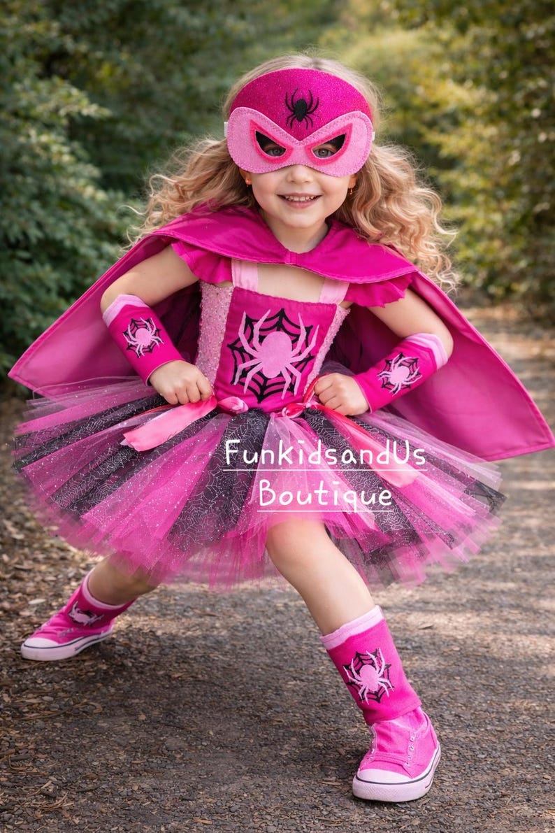 Op de afbeelding: Een roze en zwart superhelden kostuum voor kinderen. De outfit bevat een masker, cape, tutu rok, armmanchetten en beenwarmers, allemaal versierd met spinnenweb- en spinnenmotieven. De tekst "FunkidsandUs Boutique" is zichtbaar.