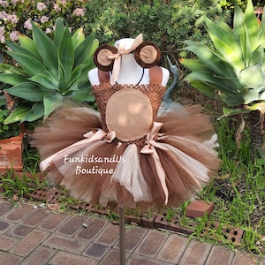 Può includere: Un abito tutu marrone e beige con fascia e orecchie abbinate. L'abito presenta un corpetto all'uncinetto e una gonna in tulle a strati. La fascia ha orecchie marroni attaccate e un fiocco. Il testo "FunkidsandUs Boutique" è visibile.