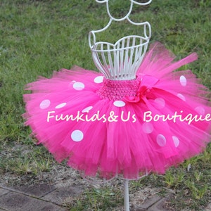 May include: A pink tulle tutu skirt with white polka dots. The skirt is on a white mannequin and has a white satin bow.