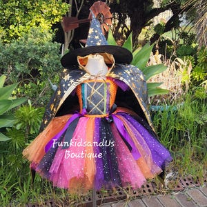 Könnte beinhalten: Ein Hexenkostüm mit schwarzem Hut und Umhang, verziert mit goldenen Sternen. Das Kleid hat einen bunten Tutu-Rock in Orange-, Rosa-, Lila- und Schwarztönen. Der Text "FunkidsandUs Boutique" ist auf dem Rock zu sehen.