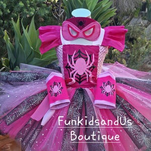 Op de afbeelding: Een roze en zwart superhelden kostuum met een spinnenthema. Het kostuum bevat een masker, top, tutu rok en armmanchetten. De top heeft een groot roze spinnendesign. De tutu rok heeft een webpatroon en is gelaagd met tule.