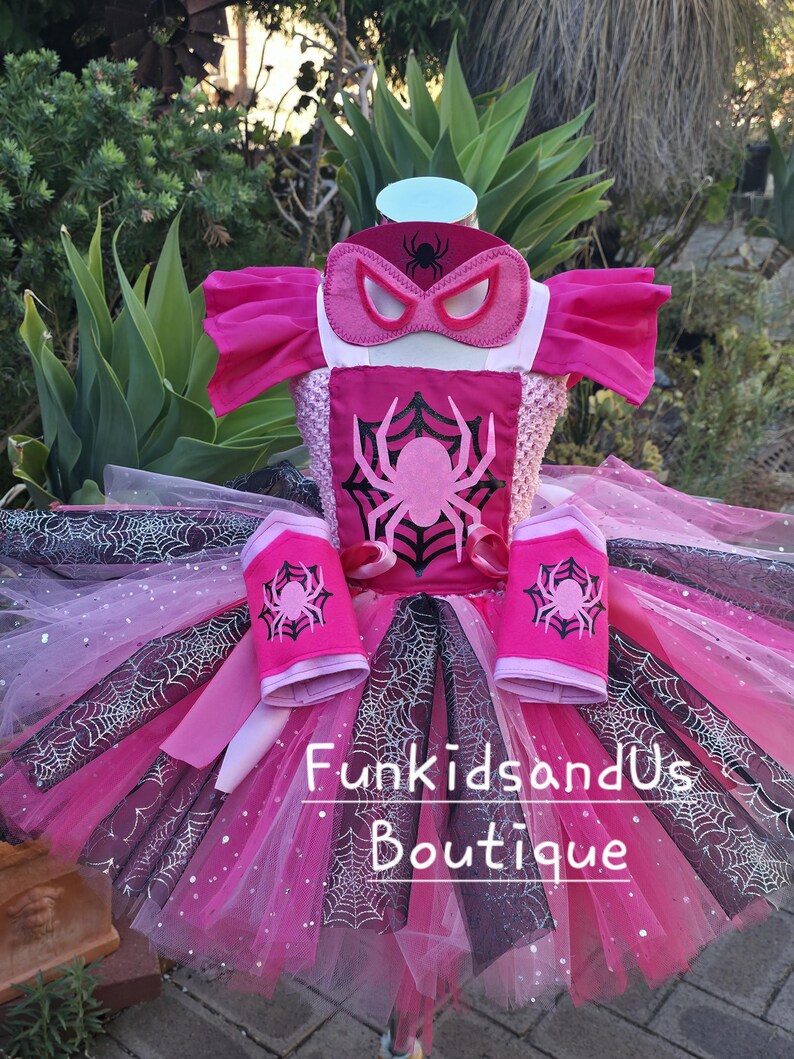 Op de afbeelding: Een roze en zwart superhelden kostuum met een spinnenthema. Het kostuum bevat een masker, top, tutu rok en armmanchetten. De top heeft een groot roze spinnendesign. De tutu rok heeft een webpatroon en is gelaagd met tule.