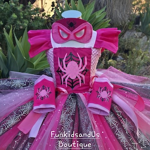 Op de afbeelding: Een roze en zwart kostuumset met spinnenthema. De set bevat een tutu-rok, een top met een spinnenembleem, armmanchetten en een masker. De tutu heeft lagen roze en zwarte tule met spinnenwebaccenten.