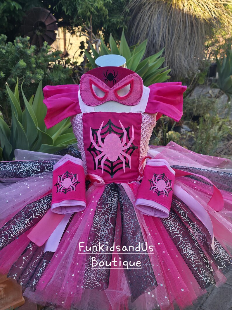 Op de afbeelding: Een roze en zwart kostuumset met spinnenthema. De set bevat een tutu-rok, een top met een spinnenembleem, armmanchetten en een masker. De tutu heeft lagen roze en zwarte tule met spinnenwebaccenten.