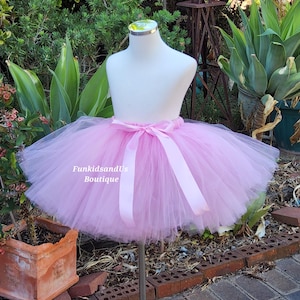 May include: A pink tulle tutu skirt with a pink satin ribbon tied around the waist. The skirt is displayed on a mannequin in a garden setting. The text "FunkidsandUs Boutique" is visible on the skirt.