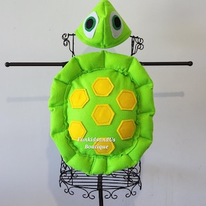 May include: A bright green turtle costume set, including a shell and a headpiece. The shell features yellow hexagon shapes and a ruffled edge. The headpiece has large white-rimmed eyes. The costume is displayed on a black metal stand. The text "FunkidsandUs Boutique" is visible.