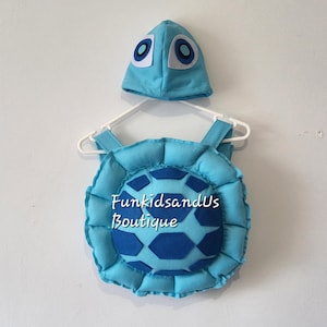 May include: A light blue turtle costume set, including a shell and a matching hat. The shell has a dark blue hexagon pattern in the center and is trimmed with a ruffled edge. The hat features large, cartoonish eyes.