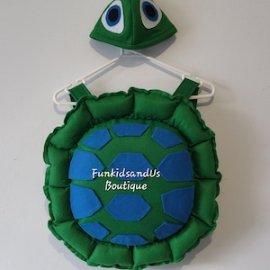 May include: A green turtle costume set, including a shell and a hat, hanging on a white hanger. The shell is green with blue geometric shapes and the text "FunkidsandUs Boutique". The hat has large blue and white eye details.