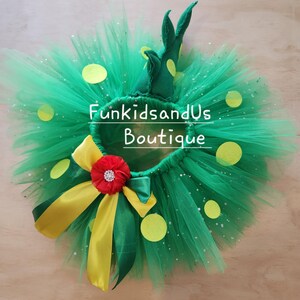 May include: A green tulle tutu skirt featuring yellow felt circles, a red fabric flower with a rhinestone centre, and yellow and green satin ribbons. A green fabric dinosaur tail is attached to the waistband. The text FunkidsandUs Boutique is visible.