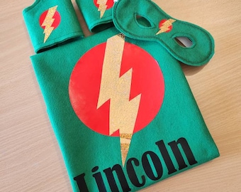 May include: Green superhero costume set with a cape, mask, and arm bands. The cape has a red circle with a gold lightning bolt and the name "Lincoln" printed below.