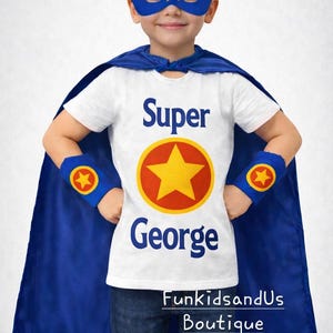 May include: A child's superhero outfit, comprising a blue cape, mask, and wrist cuffs. The white t-shirt displays the word "Super" above a yellow star emblem and the name "George". The outfit is completed with blue jeans.