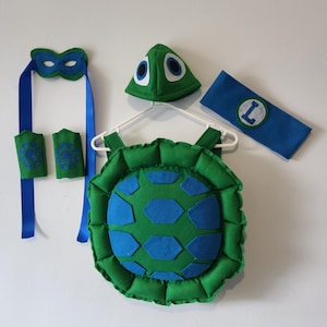 May include: A green and blue turtle costume set, including a shell, headpiece, eye mask, armbands, and a blue headband with a white and green "L" patch. The costume is made of felt and is designed for children.