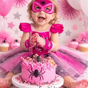 Op de afbeelding: Een kind met een roze superheldenmasker en -kostuum, voor een roze taart versierd met een zwarte spin. De taart staat op een witte taartstandaard. Roze en witte ballonnen en decoraties zijn op de achtergrond. De tekst "FunkidsandUs Boutique" is zichtbaar.