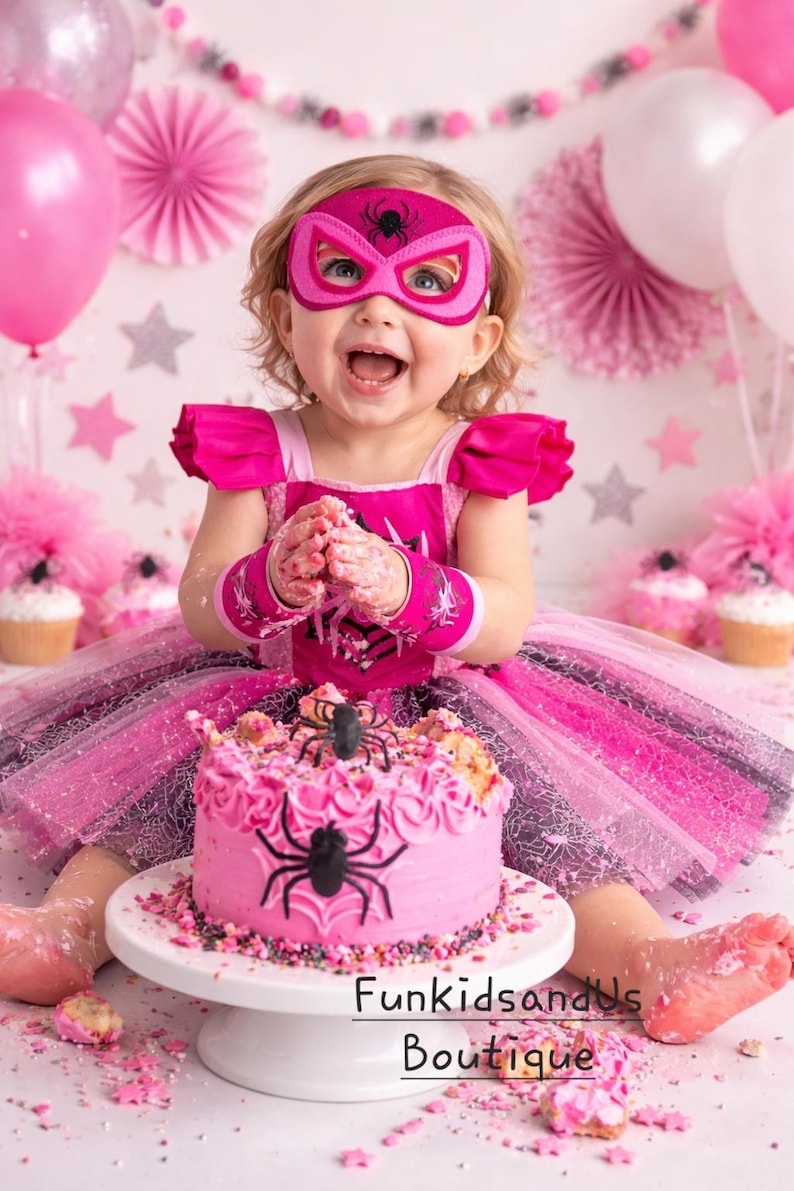 Op de afbeelding: Een kind met een roze superheldenmasker en -kostuum, voor een roze taart versierd met een zwarte spin. De taart staat op een witte taartstandaard. Roze en witte ballonnen en decoraties zijn op de achtergrond. De tekst "FunkidsandUs Boutique" is zichtbaar.