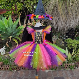 May include: A colorful witch costume with a black hat and a multi-colored tutu skirt. The skirt features layers of orange, pink, purple, and green tulle. The hat is decorated with orange and purple ribbons and flowers. The text "FunkidsandUs Boutique" is visible.