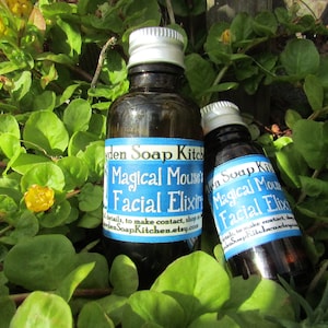 May include: Two dark glass bottles with silver caps, labelled "Magical Mouse's Facial Elixir," sit amongst green foliage and small yellow flowers. The labels are blue and white, with text that includes "den Soap Kitchen."