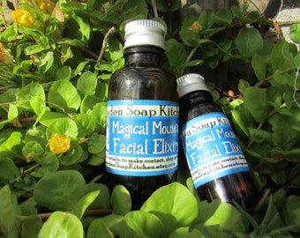 May include: Two dark glass bottles with silver caps, labeled "Magical Mouse's Facial Elixir," rest among green foliage with small yellow flowers. The labels are blue and white, with text that includes "den Soap Kitchen."