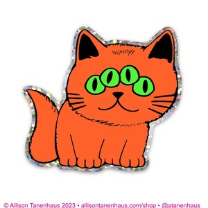 以下が含まれることがあります： 白い背景に、3つの緑色の目をしたオレンジ色の猫が座っている。