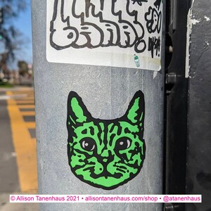 May include: A green cat sticker with a white background. The cat has a striped pattern and is looking directly at the viewer.