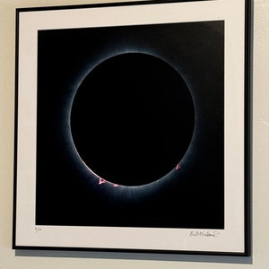 May include: A black and white photograph of a solar eclipse. The image shows the sun's corona, which is the outer atmosphere of the sun, as a bright ring around the dark disk of the moon. The image is signed "Bill Westcott" and numbered "2/20".