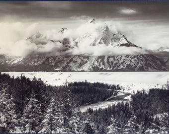 May include: A black and white photograph of a snow-capped mountain range with a lake in the foreground. The mountains are covered in clouds and snow, and the lake is calm and still. The trees in the foreground are bare and the ground is covered in snow.