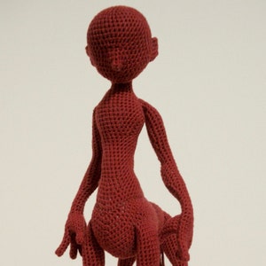 May include: A crocheted doll with a red body and no facial features. The doll is standing with its arms outstretched.