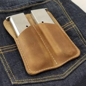 Może przedstawiać: Brązowe skórzane etui na magazynki z dwoma srebrnymi magazynkami, w tylnej kieszeni niebieskich dżinsów. Etui jest zszyte białą nicią. Dżinsy są ciemnoniebieskie.