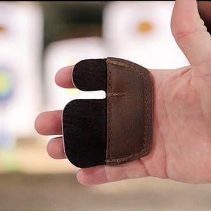 May include: A brown and black archery finger tab. The finger tab is made of leather and has a split design to protect the fingers while shooting a bow. The tab is held in a hand.
