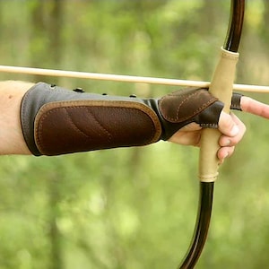 May include: A brown leather archery arm guard and finger tab are visible, along with a bow and arrow. The arm guard is designed to protect the forearm from the bowstring. The bow is held in the hand, ready to shoot the arrow.
