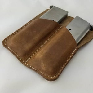 May include: A brown leather magazine pouch with two silver magazines. The pouch is stitched with white thread and has a simple, functional design. The leather has a natural, worn appearance.