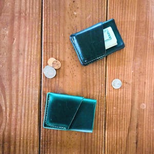 May include: Two dark green leather card holders with visible white stitching. One card holder contains a folded twenty-dollar bill. Three silver-colored coins and one copper-colored coin are positioned near the card holders.