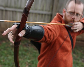 May include: A person is holding a wooden bow and arrow, aiming at a target. The bow is brown with a leather grip. The arrow is made of wood with a metal tip. The person is wearing a brown leather glove on their left hand.