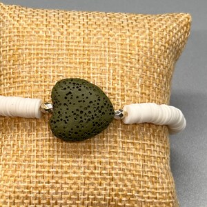 May include: A white beaded bracelet with a heart-shaped, olive-green stone at the center. The bracelet features small, flat, white beads and silver-colored accents. The heart-shaped stone has a textured surface and is the focal point of the bracelet.