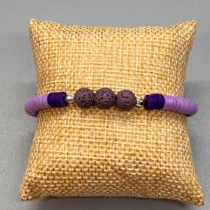 May include: A purple bracelet featuring flat, disc-shaped beads and three dark purple lava rock beads. The bracelet is accented with silver-colored spacers and is displayed on a small, textured, tan pillow.