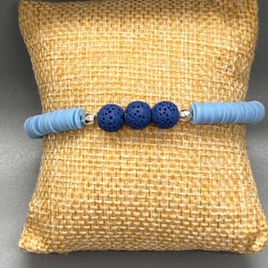 May include: A light blue beaded bracelet featuring three dark blue lava beads and silver-colored spacers. The bracelet is made with flat, disc-shaped beads and is displayed on a textured, tan surface.