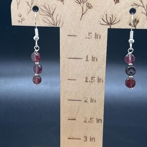 May include: Dangle earrings with three dark red, round beads on each earring. The beads are strung on a silver-colored wire. The earrings are displayed on a wooden stand with floral designs and inch measurements.