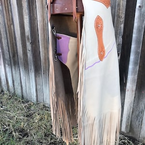 May include: A pair of white leather chaps with brown leather fringe and a brown leather belt with floral tooling. The chaps have a purple trim and a brown leather patch on the back.