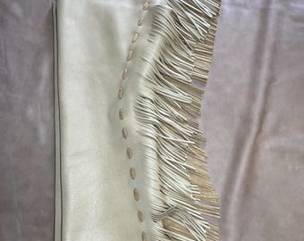 May include: Cream-colored leather chaps with long fringe along the sides. The chaps have decorative stitching and are paired with a brown leather belt with a silver buckle. The chaps are designed for horseback riding or Western wear.