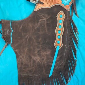 May include: Black leather chaps with turquoise fringe and stitching. The chaps have a decorative floral design on the top with a woven pattern and a turquoise flower.