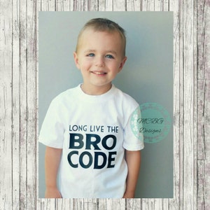 Może przedstawiać: Biała koszulka z czarnym napisem "Long Live the Bro Code". Młody chłopiec nosi koszulkę i uśmiecha się do kamery.